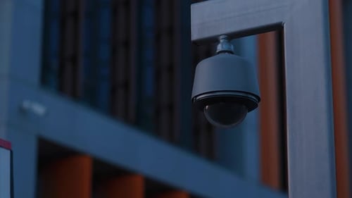 The Silhouette of a Round Black CCTV Camera Mounted on a Pole Against a Dark Corporate Building