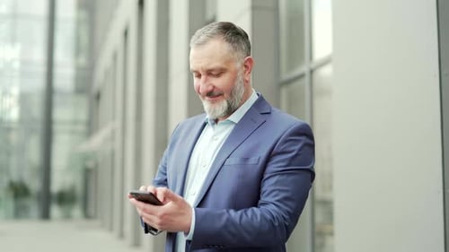 Happy mature business man in formal suit using smartphone while standing outside near the office