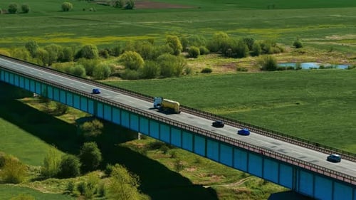 Cargo Truck and Cars Driving Across Bridge Through Green Countryside During Transport Freight Hauler