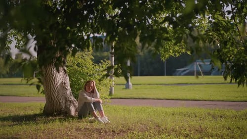 Thoughtful Woman Sitting Under Tree with Sunlight and Stadium in Background