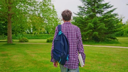 Cute Curly Teenage Boy with Headphones and a Backpack Walking Through the Park From School View From
