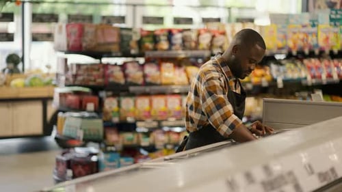 a Man with Black Skin Color in a Plaid Shirt Sorts Products in a Supermarket Refrigerator Video