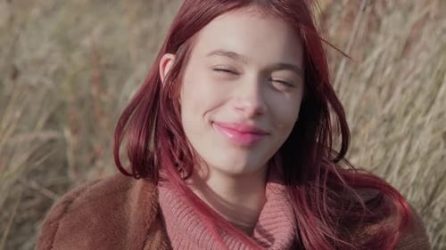 Teen girl smiling shyly while adjusting her hair, dressed warmly in a cozy autumn landscape.