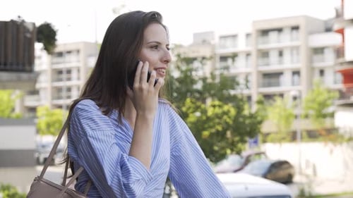 A Beautiful Young Caucasian Woman Talks on a Smartphone with a Smile in a Street in an Urban Area