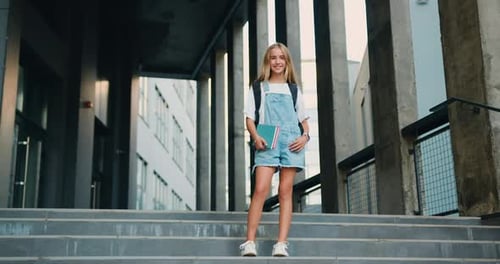 Cheerful girl hugging books posing with backpack near school building outdoor, smiling to camera.