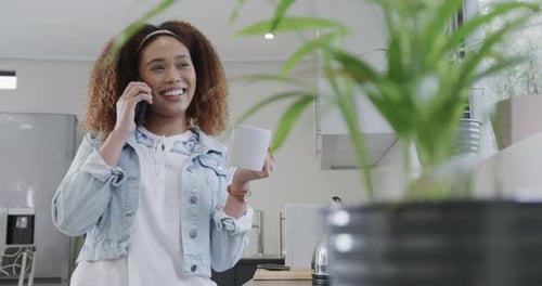 Happy biracial woman talking on smartphone, laughing and holding coffee in kitchen, slow motion