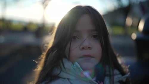 Thoughtful small girl close-up face with sunset backlight standing in playground park looking at