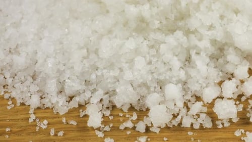 Large crystals of natural sea salt fall onto a wooden surface. A big pile of salt, lots of salt.