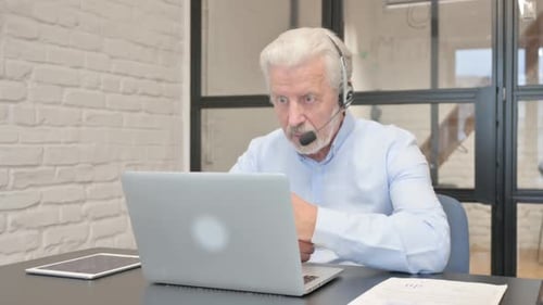 Senior Man with Headset Doing Video Chat on Laptop in Call Center