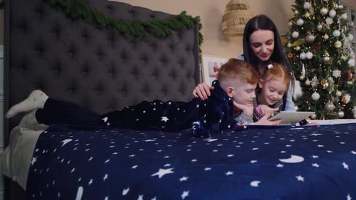 Mãe e filhos se deitam na cama e brincam no tablet