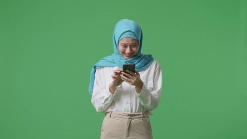 Asian Muslim Woman Smiling And Using Smartphone While Standing In The Green Screen Background