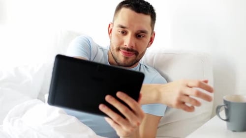 Man in bed using tablet, drinking morning beverage