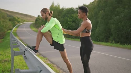 Multiethnische Paarsportler, männlich und weiblich, dehnen Beinmuskeln und trainieren auf einer leeren Landstraße