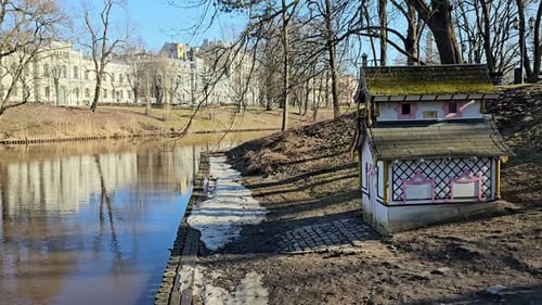 A small, white house with a red door and a blue roof. It is located on the edge of a green park, nex