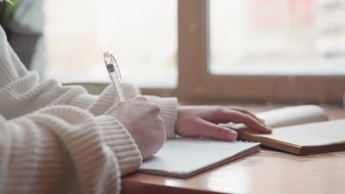 Close Up Hand Writing with Pen in Cozy Sweater Near Open Book on Wooden Table