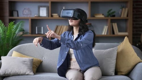 Young adult woman works using VR glasses in virtual reality simulator sitting on sofa in living room