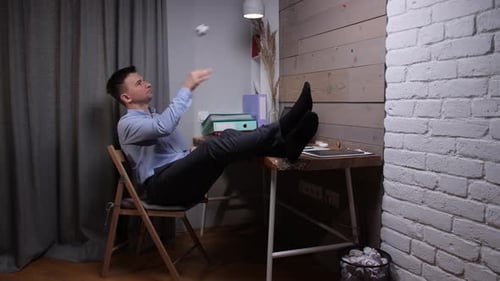 Young Man in a Home Office Sits with Feet on Desk Procrastinating Work By Tossing a Crumpled Paper
