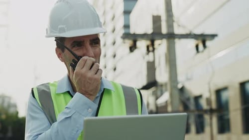 Portrait of Engineer Talking Via Radio Set Holding Laptop Standing Against Industrial Building