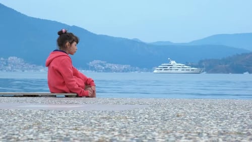 Girl Sits at Dock Overlooking Ocean and Yacht