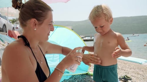 Junge Mutter trägt Sonnenschutzspray auf ihren kleinen Sohn auf, während sie an einem sonnigen Tag am Meer und Strand entspannt