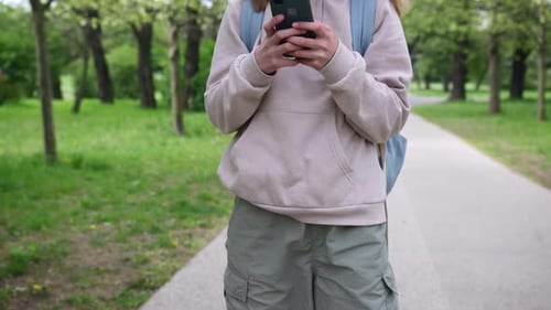 Cute Little Schoolgirl Walking And Using Smartphone In Park