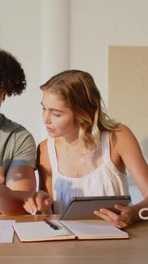 Vertical video: Diverse friends handing page, swiping tablet, toasting mugs while studying at table