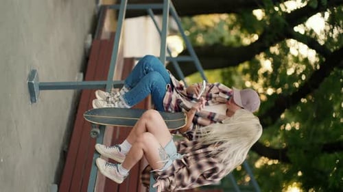 Vertical Video Two Girls in Plaid Shirts Sit and Chat on the Fence of a Skate Park in Summer Two