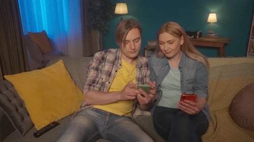 A Couple Sits in the Evening on the Sofa in the Living Room with Phones Man and Woman Choose a Movie