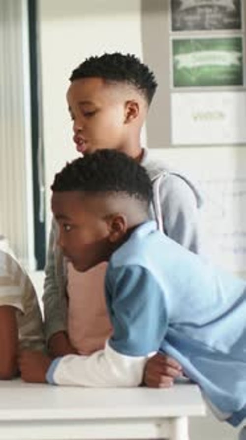 Vertical video: Leaning two boys observing left task at white table, noting grey hoodie
