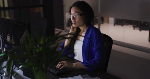 Video of tired asian businesswoman in glasses sitting at desk working at night in office
