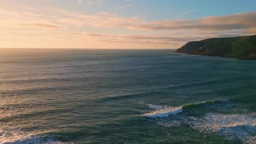 Tranquil ocean swell at dusk washing green hills seashore in slow motion aerial view