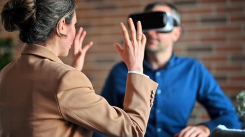 Man using a Virtual Reality headset in an office while woman explains and gesticulates