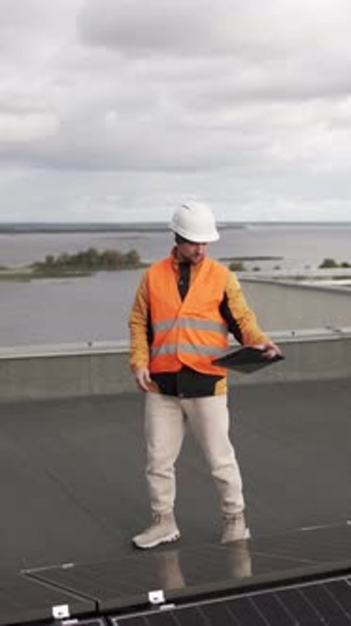 An engineer in protective gear holds a tablet near solar panels during a roof inspection.