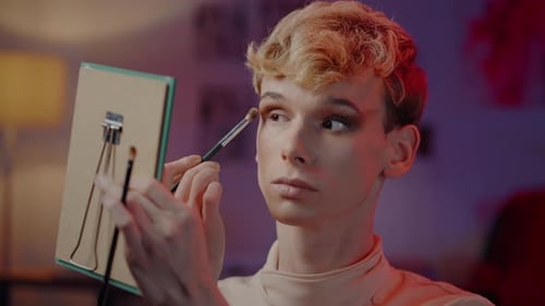 Front View of a Non Binary Male in Female Clothes Applying Makeup in Front of Cosmetic Mirror