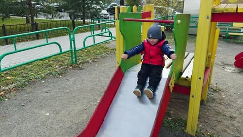Joyful child sits on slide tapping feet. Toddler boy in red jacket on the playground.