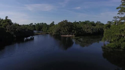 Aerial shot of a lake on a sunny day. People walk near a pavilion in the background. Camera moves do