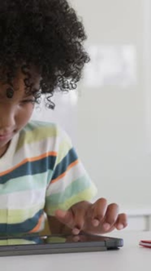 Vertical video: Leaning forward, youth in striped tee tapping tablet studying at home, with red pen