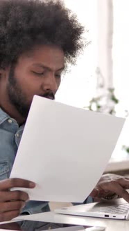 Man Reads Documents and Types on Laptop Indoors