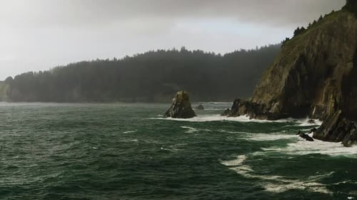 Rough waters of Pacific Ocean crash against rocky Oregon coastline, foggy day.