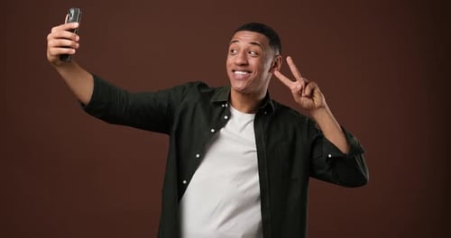 Smiling Young African American Man Making Selfies on Phone Standing Over Brown Studio Background