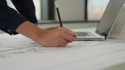 Architect Writing on Blueprint with Laptop in Office