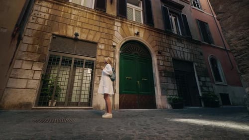 Young Woman in a White Dress and Hat with a Backpack Looking Up at an Old Stone Building with Green