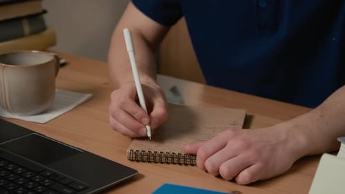 Mann schreibt in Notizbuch, während er am Schreibtisch für Hausaufgaben am College studiert