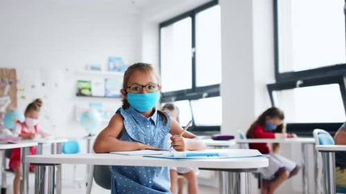 Elementary school girl wearing face mask writes at desk in classroom
