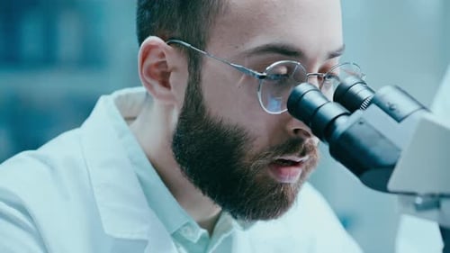 Scientist Looking through Microscope and Talking to Assistant in Laboratory