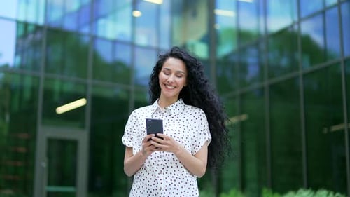Young smiling female employee is using a smartphone while walking on street near an office building.