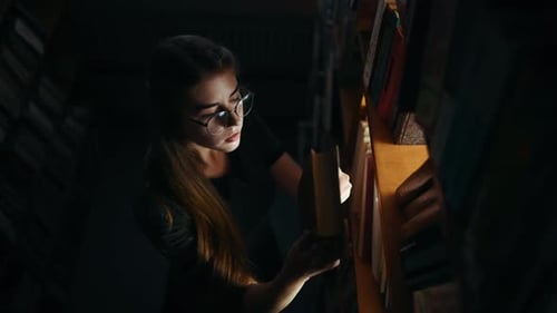 Girl in eyewear searching for the book in dark room with light beam comes through the shelves