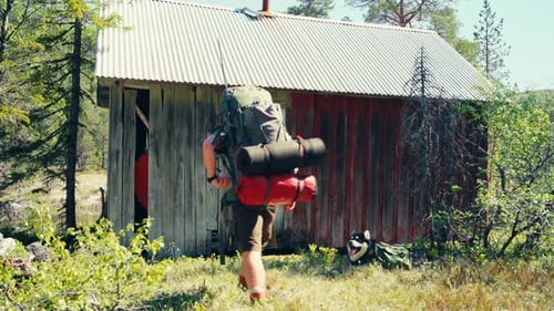 Man With Backpack Resting With Dog Near Small Wooden Hut In The Mountain.