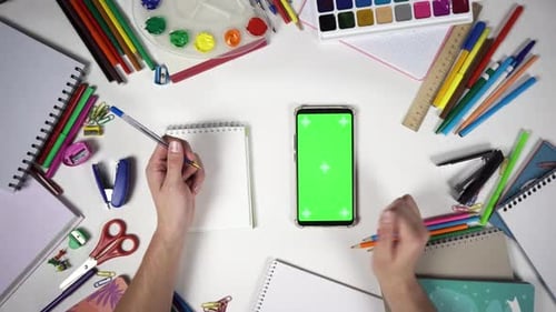 Left Handed Man Makes Notes and Swipes on the Smartphone with a Green Screen Lying on the Desk