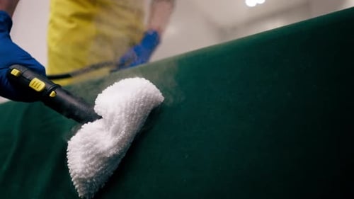 Close Up in an Apartment Cleaner in a Yellow Apron Does a Steam Cleaning of a Green Sofa with